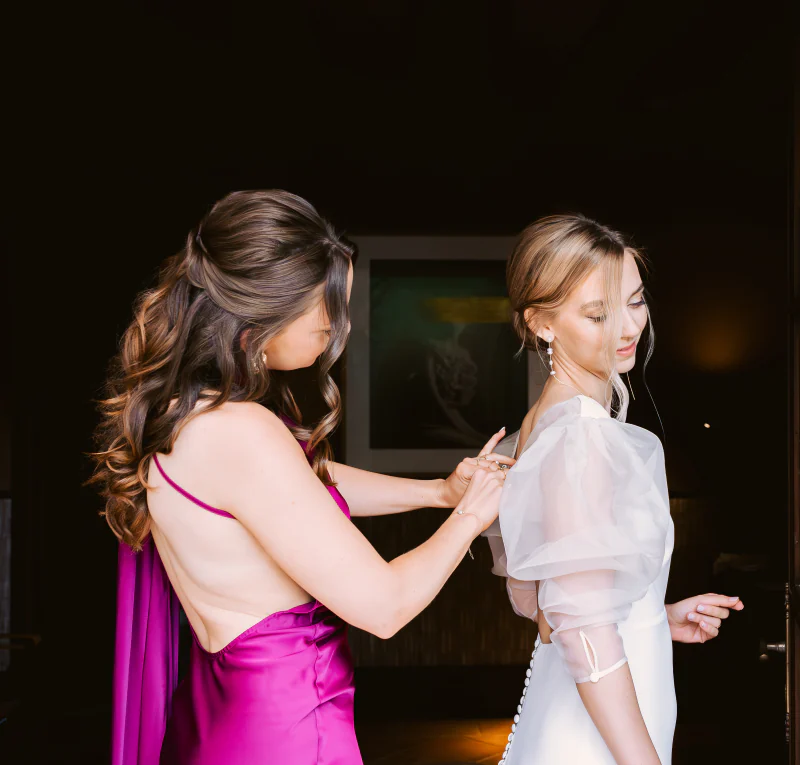 A bridesmaid helping the bride with the buttons of her dress before the ceremony