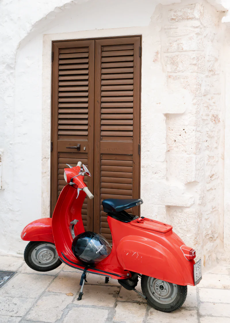 A red Vespa parked against a white wall with wooden shutters