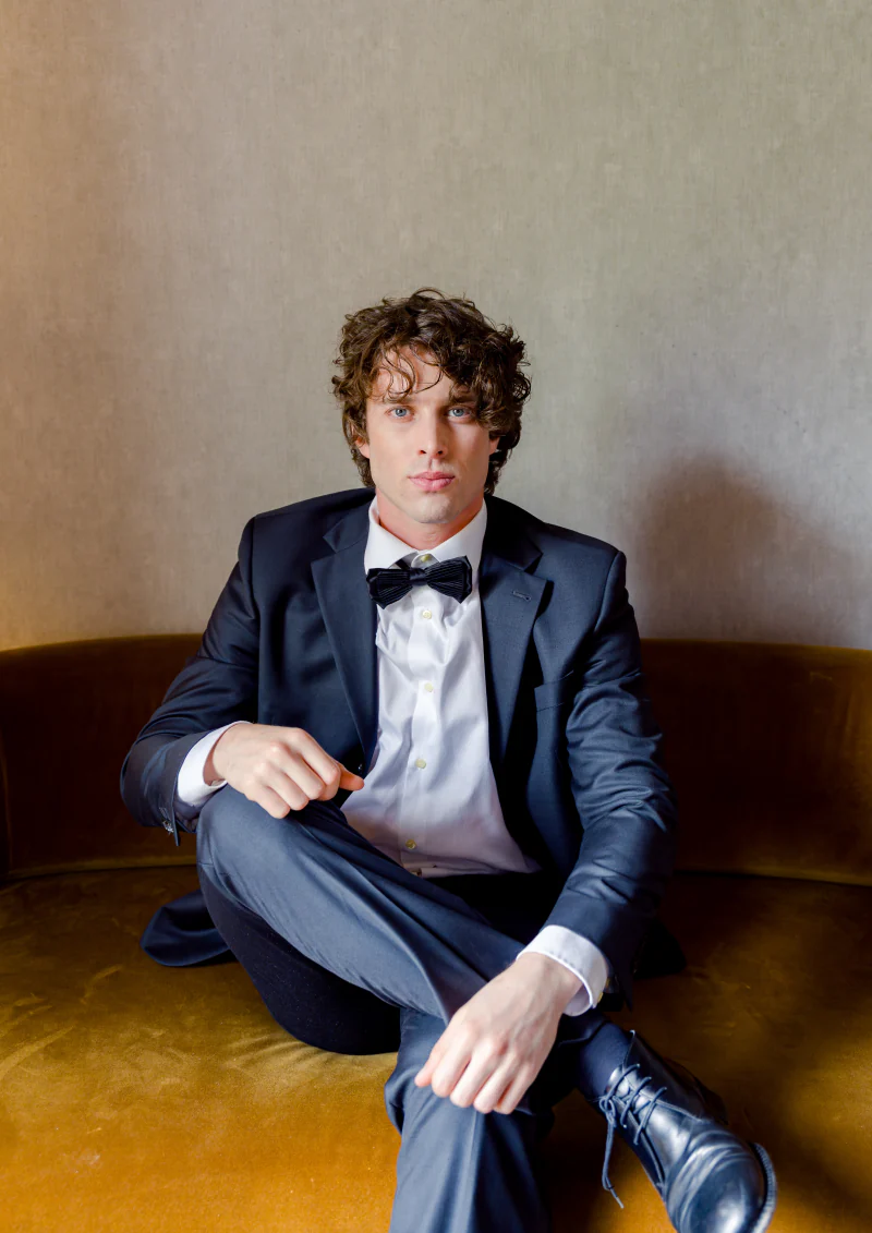 Groom in a navy tuxedo seated on a velvet sofa