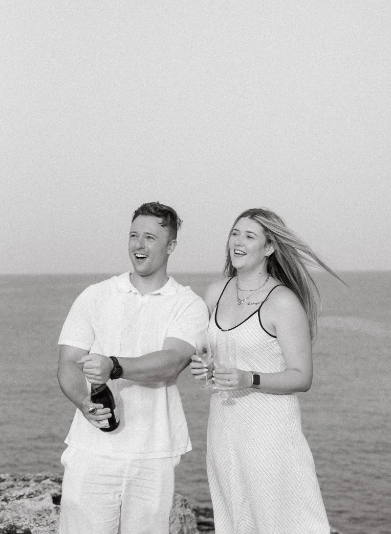 Black-and-white: a couple laughing as champagne is opened by the sea